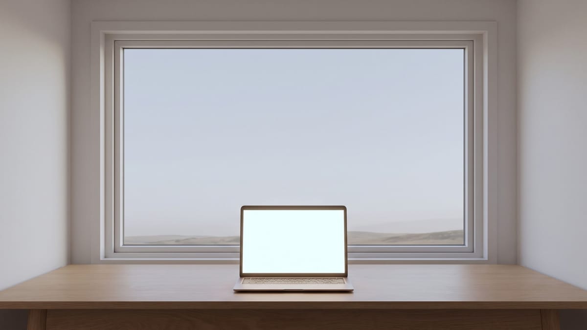Un computer portatile aperto al centro di una scrivania vuota davanti a una finestra, con la luce del mattino